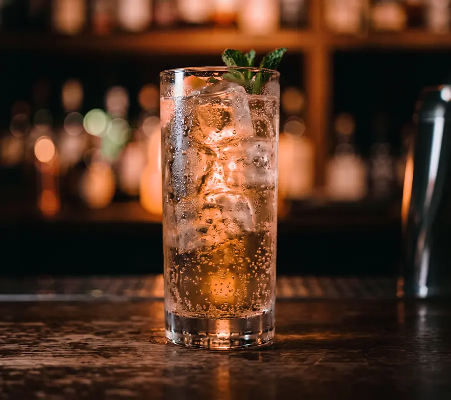 Ein Longdrinkglas mit Eiswürfeln, prickelndem Getränk und Minzzweig steht auf einer Theke vor unscharfem Hintergrund.